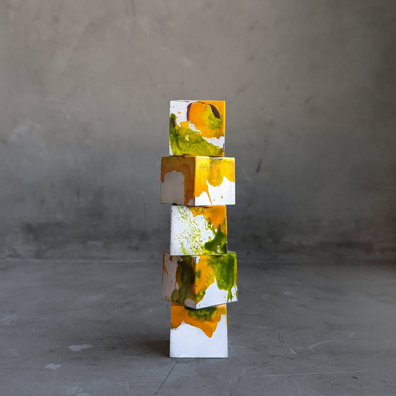 Five gourmet chocolate truffles in white, yellow, and green artfully stacked on top of one another in front of a concrete background.